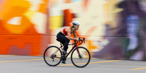 Woman cycling at high speed in front of a colorful graffiti wall, wearing athletic gear and a helmet.