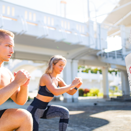  A man and woman exercising outdoors with whey protein and pre-workout supplement containers in the foreground. 